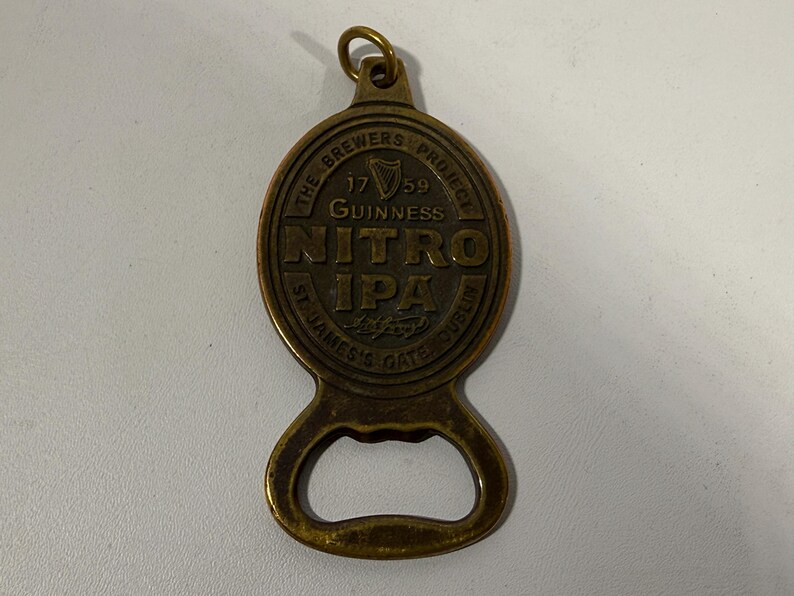 May include: A brass-colored bottle opener with a circular top featuring the text "NITRO IPA" and "GUINNESS" with the year "1759". The opener has a loop at the top and a bottle cap opener at the bottom.