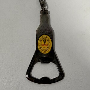 May include: A metal bottle opener in the shape of a Guinness Extra Stout bottle. The opener has a yellow and black label with the Guinness logo and text. A small chain and clasp are attached to the top.
