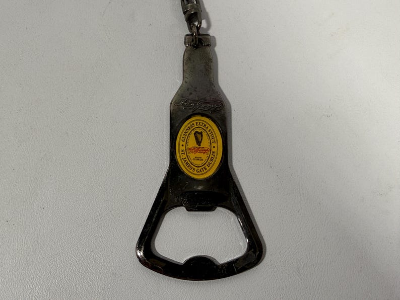 May include: A metal bottle opener in the shape of a Guinness Extra Stout bottle. The opener has a yellow and black label with the Guinness logo and text. A small chain and clasp are attached to the top.