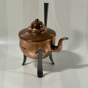 May include: A small copper teapot with a black handle and three black legs. The teapot has a round body and a lid with a knob. The teapot is inscribed with the text "Klatt".