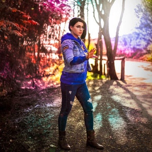 Può includere: Una donna con una giacca di jeans blu, una felpa blu, jeans blu e stivali neri si trova in un ambiente boschivo. Lo sfondo è una sfocatura di alberi colorati e luce solare.