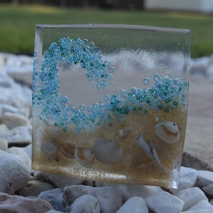 May include: A clear resin block with a wave of blue and green beads, sand, and seashells. The block is sitting on a bed of white rocks.