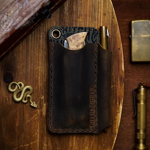 May include: A dark brown leather pocket organizer with the embossed text HITCH AND TIMBER, containing a small folding knife with a light wood handle and a brass pen. The organizer features a metal grommet at the top corner and visible stitching.