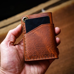 May include: A brown leather wallet with a card in the front pocket. The wallet has a brass grommet and the words "Hitch + Timber" are embossed on the front. The wallet is held in a hand.