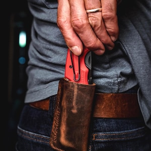 Könnte beinhalten: Ein rotes und schwarzes Klappmesser mit Taschenclip ist teilweise in einer braunen Lederscheide zu sehen. Die Scheide ist an einem Gürtel befestigt. Das Messer wird von einer Person gehalten, die ein graues Hemd und eine blaue Jeans trägt. Auf der Scheide steht der Text "HITCHHIKER".