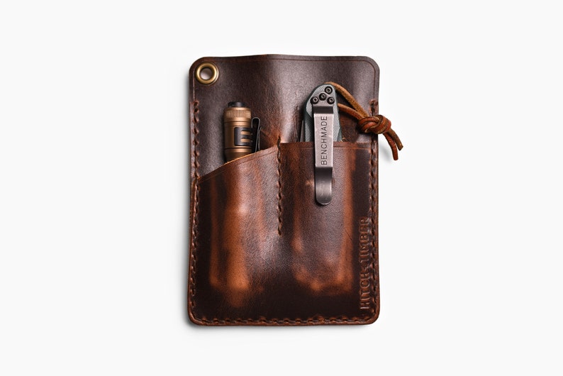 Peut inclure: Un organiseur de poche en cuir marron avec deux poches, une pour un stylo et une pour un couteau pliant. Le couteau est de la marque Benchmade et l'organisateur est &eacute;tiquet&eacute; "Hitch & Tiger".