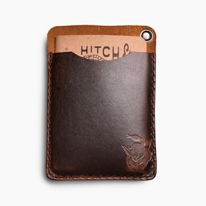 Peut inclure: Un porte-cartes en cuir marron avec un bord cousu et une petite boucle en m&eacute;tal. Le porte-cartes a une poche sur le devant et un petit motif en relief d'un serpent dans le coin inf&eacute;rieur droit. Le texte "HITCH" est visible sur un morceau de papier &agrave; l'int&eacute;rieur de la poche.