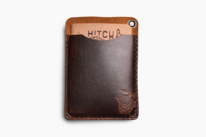 Peut inclure: Un porte-cartes en cuir marron avec un bord cousu et une petite boucle en m&eacute;tal. Le porte-cartes a une poche sur le devant et un petit motif en relief d'un serpent dans le coin inf&eacute;rieur droit. Le texte "HITCH" est visible sur un morceau de papier &agrave; l'int&eacute;rieur de la poche.