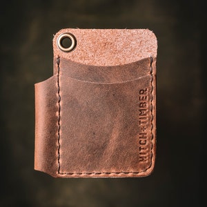 May include: A brown leather wallet with a single pocket and a brass ring. The wallet has the text "HITCH + TIMBER" embossed on the side.