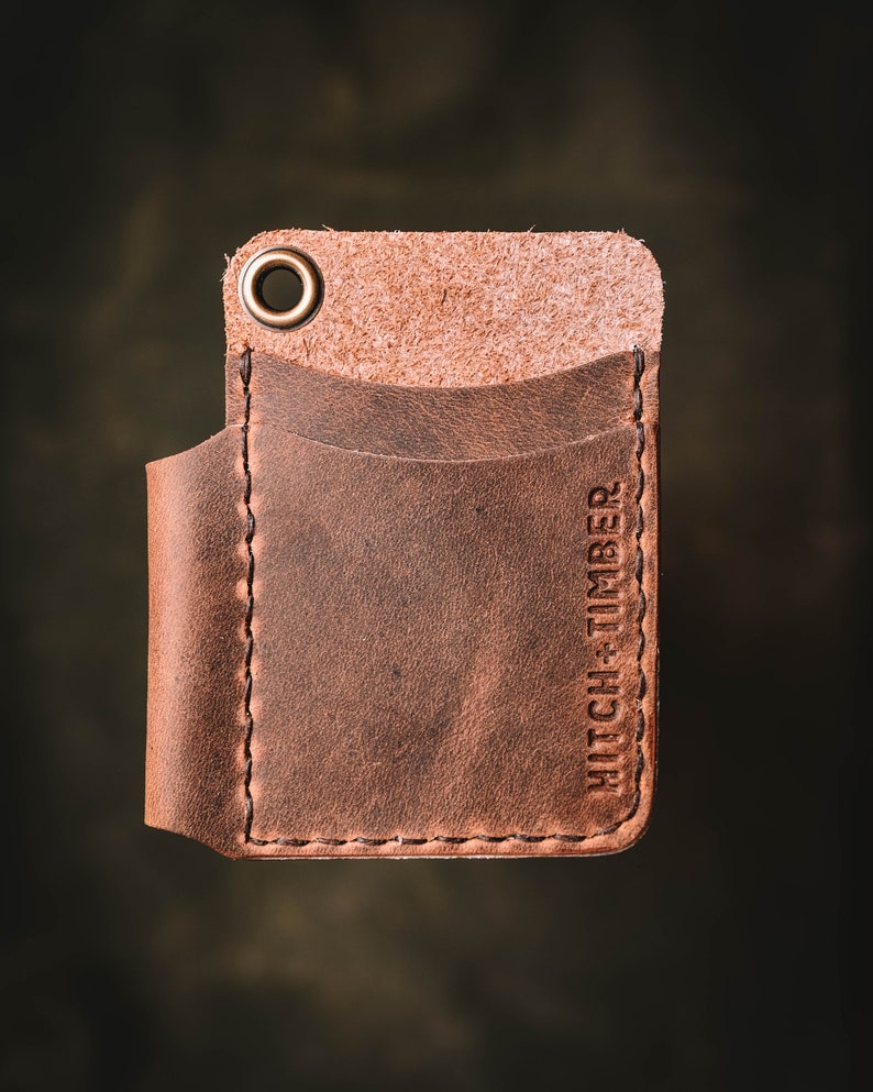 May include: A brown leather wallet with a single pocket and a brass ring. The wallet has the text "HITCH + TIMBER" embossed on the side.