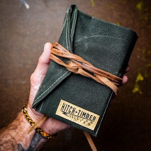 Puede incluir: Una cartera de lona verde oscuro con una solapa plegable asegurada por un fino cordón de cuero marrón envuelto alrededor de un ojal metálico. Una etiqueta rectangular beige en el frente presenta el texto negro HITCH + TIMBER y AMERICAN CRAFTED.