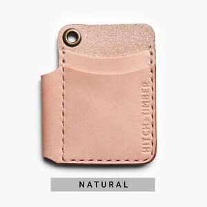 May include: A light pink leather card holder with a stitched edge and a brass grommet. The card holder has a single pocket and the text "HITCH+TIMBER" embossed on the side. The card holder is labeled "NATURAL".