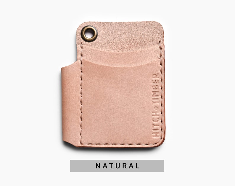 May include: A light pink leather card holder with a stitched edge and a brass grommet. The card holder has a single pocket and the text "HITCH+TIMBER" embossed on the side. The card holder is labeled "NATURAL".