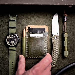 May include: A green leather wallet with a card slot and a pocket for cash. The wallet is sitting on a green canvas background with a watch, a knife, and a flashlight.