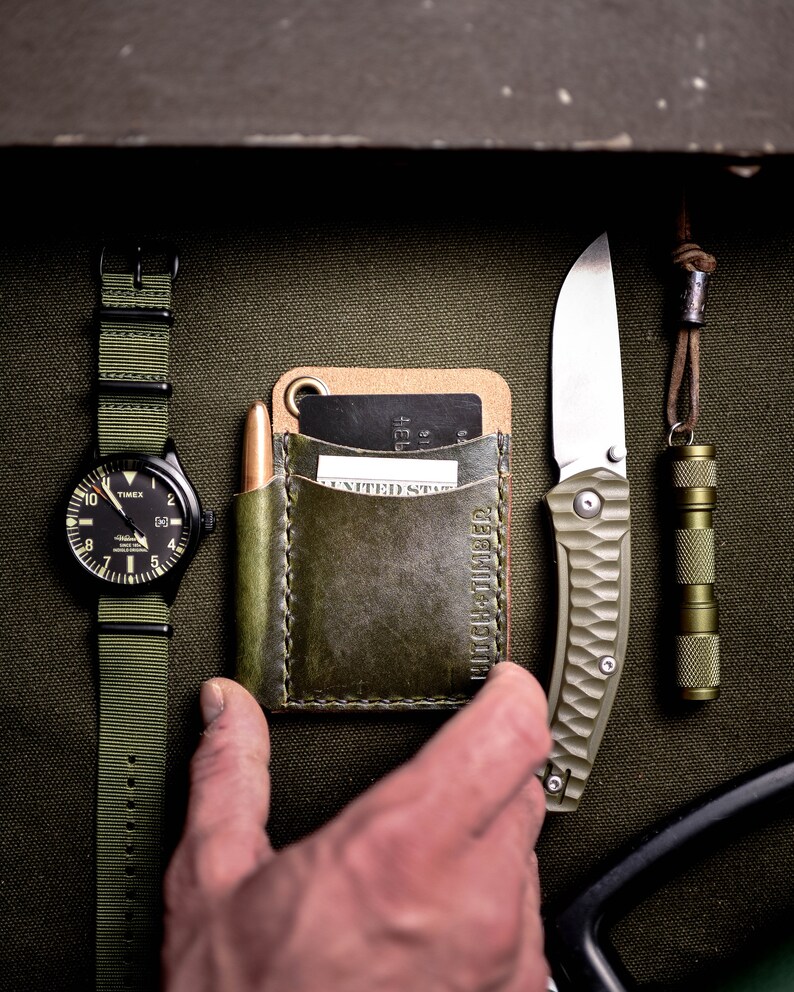 May include: A green leather wallet with a card slot and a pocket for cash. The wallet is sitting on a green canvas background with a watch, a knife, and a flashlight.