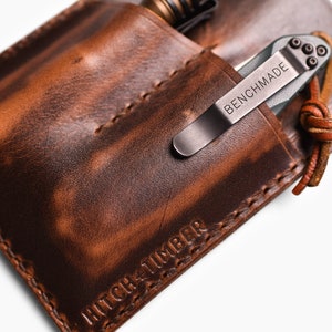 Peut inclure: Un porte-couteau de poche en cuir marron avec un bord cousu et le texte "HITCH & TIMBER" en relief sur le fond. Un couteau de poche argent&eacute; avec le texte "BENCHMADE" est &agrave; l'int&eacute;rieur du porte-couteau.