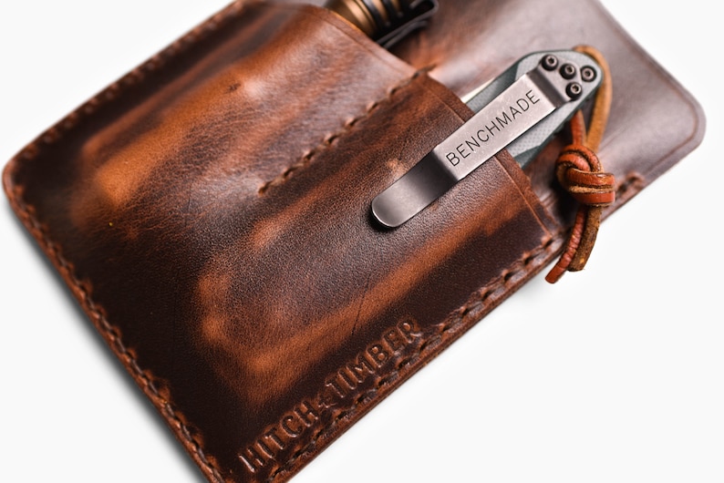 Peut inclure: Un porte-couteau de poche en cuir marron avec un bord cousu et le texte "HITCH & TIMBER" en relief sur le fond. Un couteau de poche argent&eacute; avec le texte "BENCHMADE" est &agrave; l'int&eacute;rieur du porte-couteau.