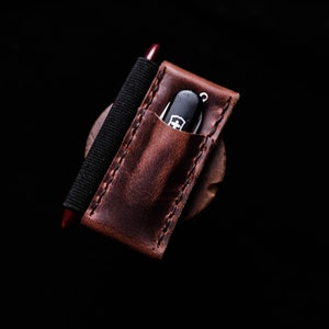 Puede incluir: Un portacuchillos de bolsillo de cuero marrón con una banda elástica negra y un cuchillo de bolsillo plateado con mango rojo.