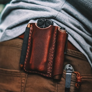 Puede incluir: Un portaherramientas multiusos de cuero marrón con el texto "HITCHHIKER" grabado en la parte delantera. Una herramienta multiusos negra es parcialmente visible dentro del portaherramientas. Una herramienta multiusos negra con el texto "BENCHMADE" está enganchada a un bucle de cinturón marrón.