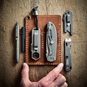 Peut inclure: Un portefeuille en cuir marron avec plusieurs poches pour transporter divers outils. Le portefeuille contient un stylo argent&eacute;, un multi-outil avec une cl&eacute; hexagonale, un petit couteau et un ouvre-bouteille. Le texte "HITCH + TIMBER" est imprim&eacute; sur le portefeuille.