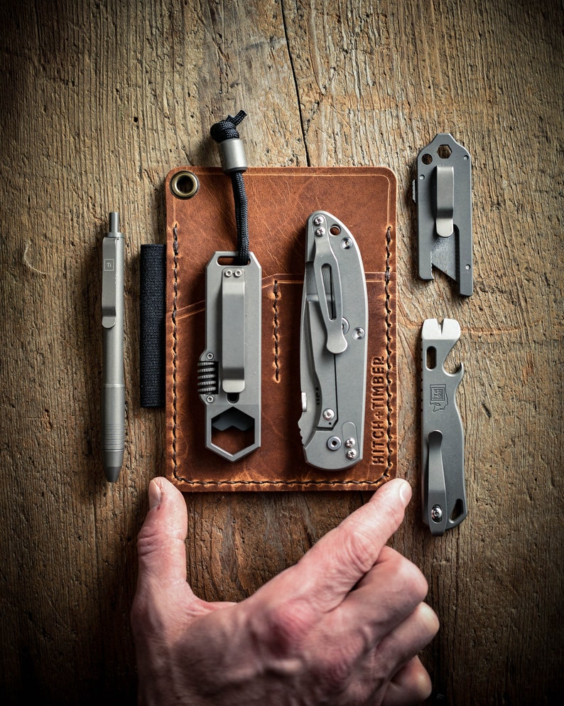 Peut inclure: Un portefeuille en cuir marron avec plusieurs poches pour transporter divers outils. Le portefeuille contient un stylo argent&eacute;, un multi-outil avec une cl&eacute; hexagonale, un petit couteau et un ouvre-bouteille. Le texte "HITCH + TIMBER" est imprim&eacute; sur le portefeuille.