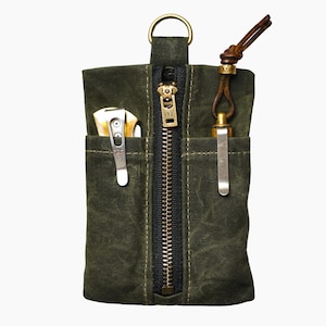 May include: An olive green waxed canvas pocket organizer featuring a central black zipper with a brass YKK pull, two exterior slip pockets, a brass D-ring at the top, and a brown leather cord attached to a brass bead on the right side.