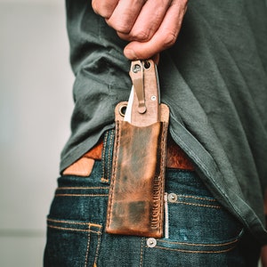 Peut inclure: Un étui à couteau en cuir marron avec un couteau de poche argenté à l'intérieur. L'étui porte le texte "Hitch & Timber" en relief sur le côté. Le couteau est rangé dans la poche avant d'un jean bleu.