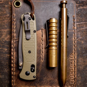 Peut inclure: Un portefeuille en cuir marron avec un bord cousu et une poche pour un couteau, un stylo et une lampe de poche. Le portefeuille porte le texte "HITCH + TIMBER" en relief sur le devant.