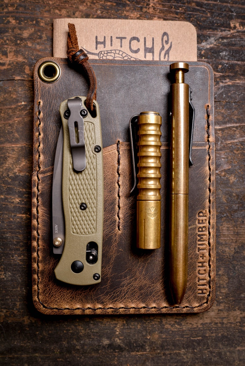 Peut inclure: Un portefeuille en cuir marron avec un bord cousu et une poche pour un couteau, un stylo et une lampe de poche. Le portefeuille porte le texte "HITCH + TIMBER" en relief sur le devant.