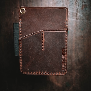 Peut inclure: Un portefeuille en cuir marron avec un bord cousu et une seule poche. Le portefeuille a un &oelig;illet en laiton et le texte "HITCH + TIMBER" en relief sur le devant.
