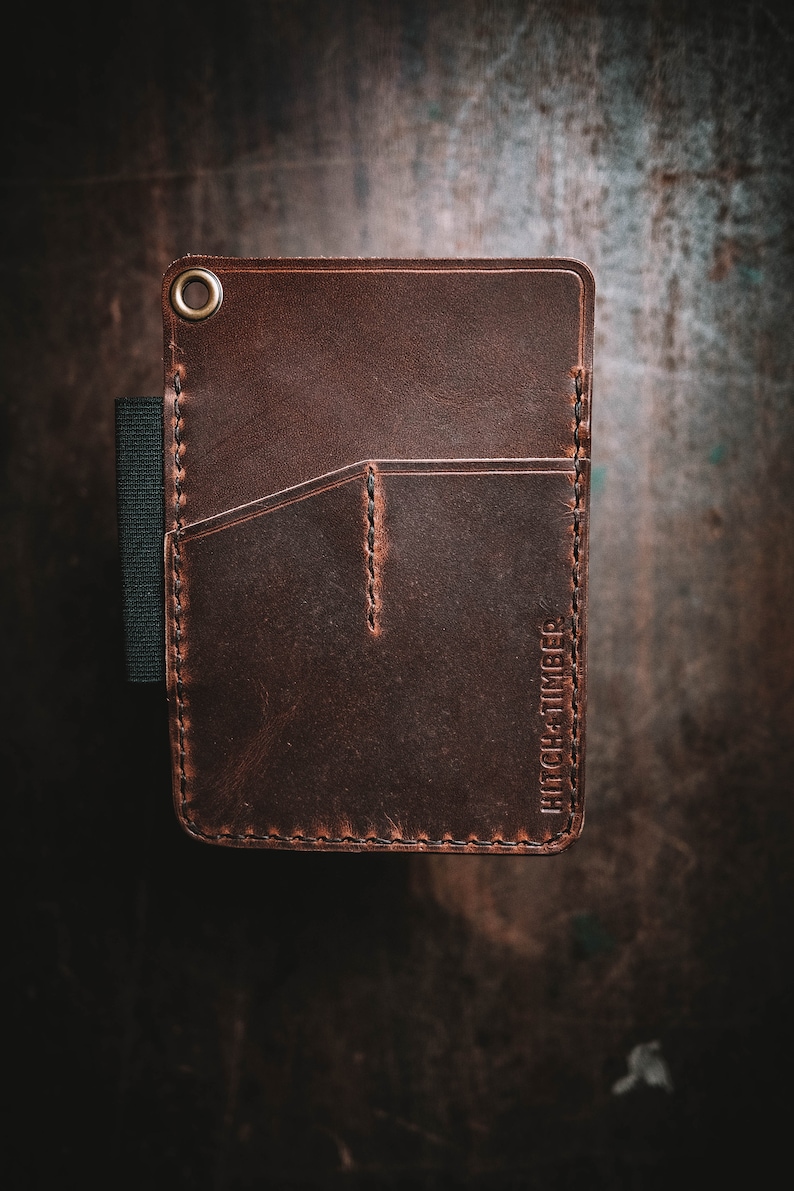 Peut inclure: Un portefeuille en cuir marron avec un bord cousu et une seule poche. Le portefeuille a un &oelig;illet en laiton et le texte "HITCH + TIMBER" en relief sur le devant.