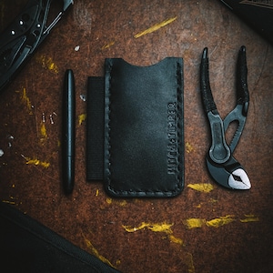 May include: A black leather cardholder with the text "HITCH+TIMBER" is centered on a brown surface. A black pen and a pair of black pliers are on either side. A black multi-tool and a small black pouch are also visible.