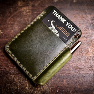 May include: A green leather wallet with a stitched edge and a card slot. The wallet has a pen holder and a card inside with the text "Thank You!" printed on it.