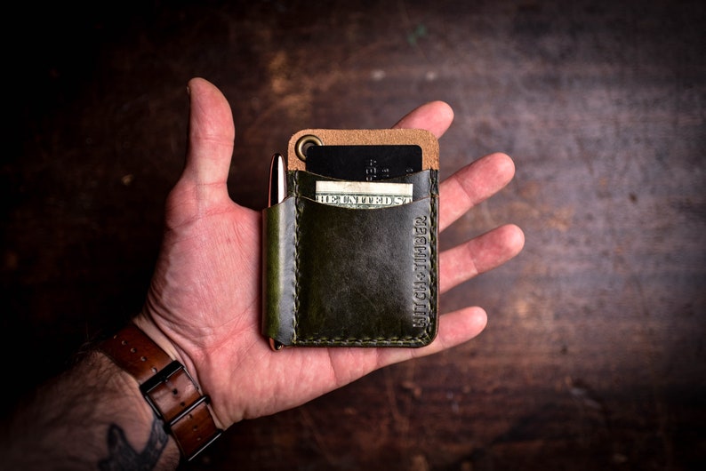 May include: A green leather wallet with a brown leather flap and a pen holder. The wallet has a card slot and a money pocket. The text "HIGH & TIBER" is embossed on the front of the wallet.