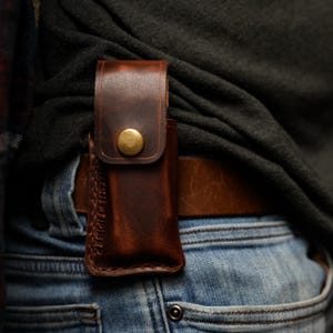 Pode incluir: Uma bainha de couro castanho com fecho de botão de latão, presa a um cinto. A bainha foi concebida para guardar uma pequena ferramenta ou faca. O couro tem um aspeto rico e texturizado, e as costuras são visíveis nas bordas.