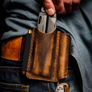 May include: A brown leather multi-tool sheath with the text "HITCH + TIMBER" embossed on the front. A silver multi-tool is partially visible in the sheath. A black pen is secured on the side with a black elastic band. The sheath is attached to a belt loop.