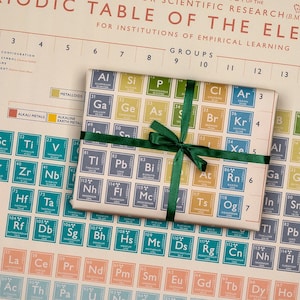 May include: A gift wrapped in paper printed with a periodic table of elements, tied with a green ribbon. The background features a larger periodic table chart. The gift wrap includes element symbols and atomic numbers.