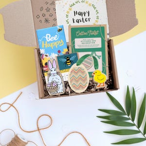 Pode incluir: Uma caixa de cartão aberta cheia de itens com tema de Páscoa. Inclui um livro "Bee Happy", um cartão "Happy Easter", uma decoração de ovo costurada, recortes de coelho e pintinho e um cartão de abelha. Sobre uma superfície branca e amarela.