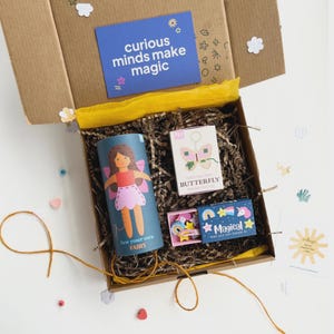 Könnte beinhalten: Ein Karton gefüllt mit Bastelsets. Darin befinden sich ein zylindrisches "Sew your own Fairy"-Set, ein "Butterfly"-Set und eine kleine Schachtel mit der Aufschrift "Magical". Eine blaue Karte mit dem Text "curious minds make magic" ist sichtbar.