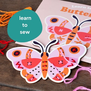 Può includere: Un artigianato in legno a forma di farfalla rosa e arancione con fori per infilare il filo. La farfalla ha un motivo rosso, bianco e arancione. Il testo "Learn to Sew" è visibile sullo sfondo.