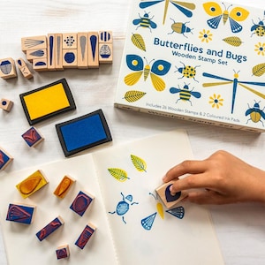 May include: A wooden stamp set with the text "Butterflies and Bugs" includes 26 stamps and 2 ink pads. The stamps feature insect and leaf designs. The ink pads are yellow and blue. A hand is using a stamp on a white notebook.