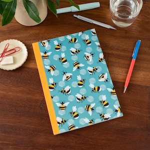 May include: A notebook with a teal cover patterned with yellow and black bees and white floral accents. The notebook has a yellow spine. Other items on the wooden surface include pens, a small dish with paperclips, and a glass of water.