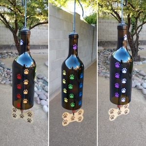May include: Three wine bottle wind chimes with paw prints in different colors. The wind chimes are made from recycled wine bottles and have a wooden bone charm at the bottom.