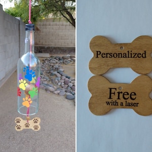 May include: A glass bottle wind chime with paw print designs in rainbow colors. The bottle is suspended from a pink string and has a wooden bone charm at the bottom. The image also shows two wooden bone-shaped tags with the text "Personalized" and "Free with a laser" on them.