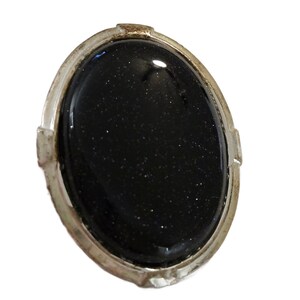 May include: Oval-shaped, dark blue gemstone set in a silver-toned metal frame. The gemstone has a speckled appearance, and the metal frame has a decorative, slightly textured design. The jewelry piece is set against a white background.