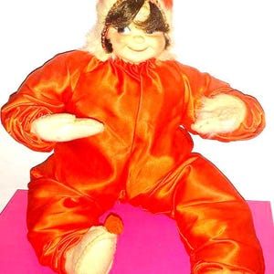 May include: A vintage doll wearing a red satin jumpsuit with a white fur trim and a matching red satin hat. The doll is sitting on a pink surface.