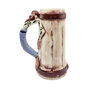 May include: A ceramic beer stein with a whimsical design. The handle is shaped like a person in a white shirt with green polka dots and blue jeans. The stein has a wood-grain appearance with brown accents.
