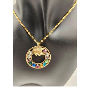 May include: Gold-toned necklace featuring a circular pendant with a leaf design and a ring of colorful gemstones. The pendant hangs from a delicate chain. The gemstones are in various colors, including blue, red, yellow, and purple.