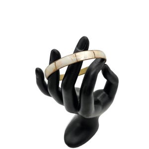 May include: A gold-toned bangle bracelet with inlaid rectangular mother-of-pearl segments. The bracelet is displayed on a black hand-shaped jewelry holder against a white background. The bracelet is a piece of jewelry.