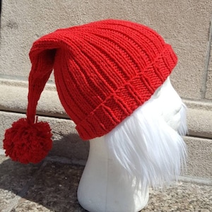 Puede incluir: Un gorro rojo de punto con una larga punta puntiaguda y un pomp&oacute;n en el extremo. El gorro est&aacute; en una cabeza de maniqu&iacute; blanca con una barba blanca.